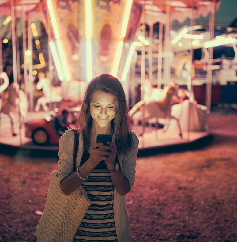 Woman at a theme park standing in front of a carousel at night looking at her mobile app