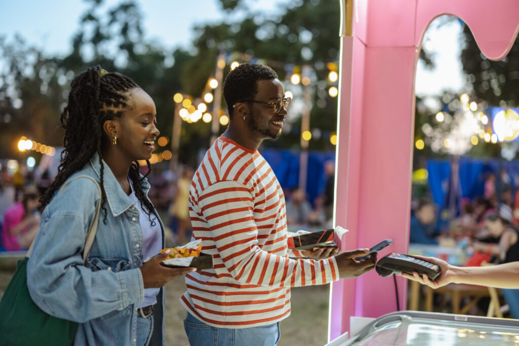Two friends at a lively outdoor fair share a smile as one pays with a mobile payment device for snacks, capturing casual fashion, warmth, and social connection in a vibrant setting.